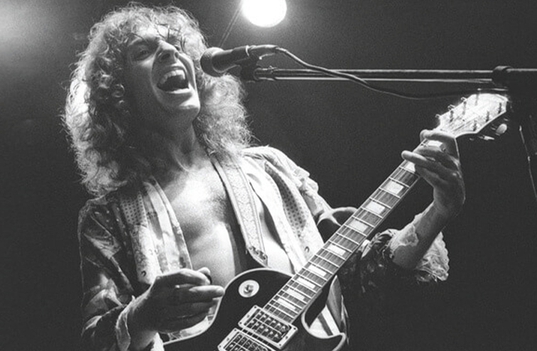 A black and white photo of a long haired Peter Frampton playing guitar and singing on stage in the 1970's.