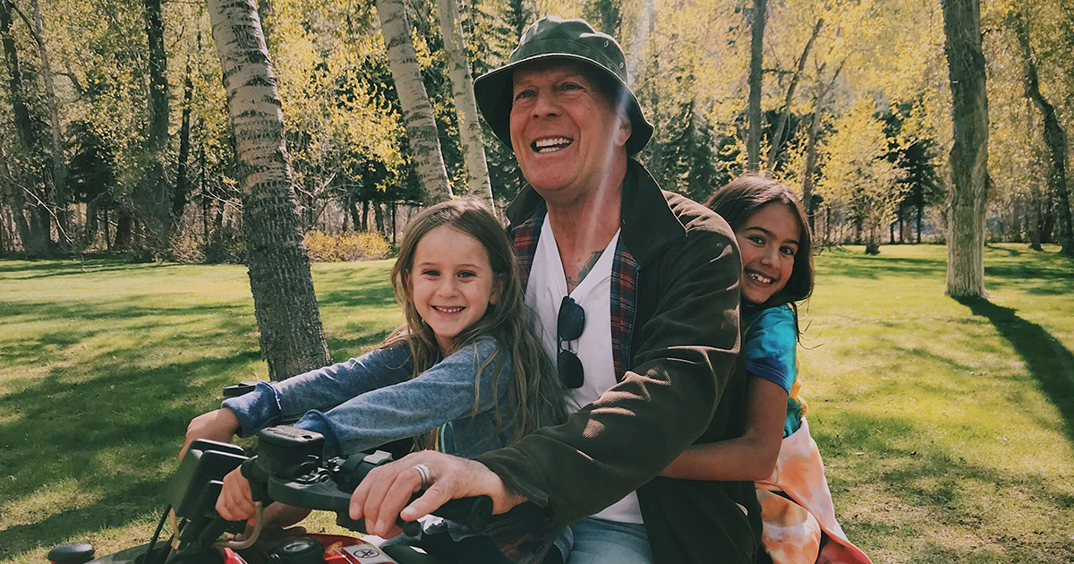 Bruce Willis rides an ATV with two of his children in a tree-lined park.
