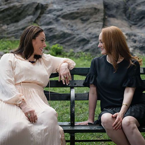 Two women sit on a park bench, smiling at each other. One wears a light, flowing cream-colored dress, and the other wears a short-sleeved black dress. A rocky hill and greenery are visible in the background.