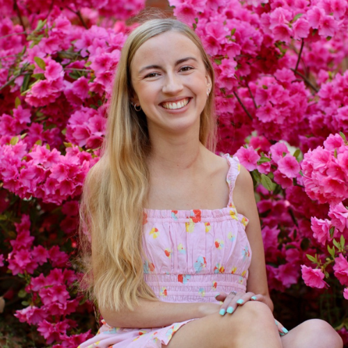  young woman with long blonde hair sits in front of vibrant pink flowers, smiling warmly. She is wearing a light pink sundress with a colorful fruit pattern and has mint green nail polish.