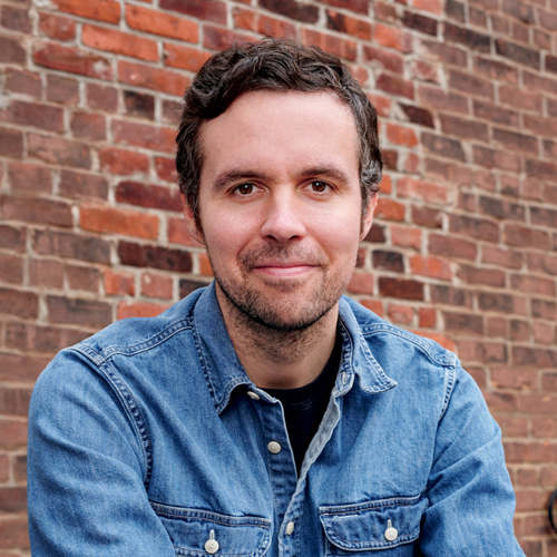 A photograph of author John Hendrickson wearing a blue denim shirt.