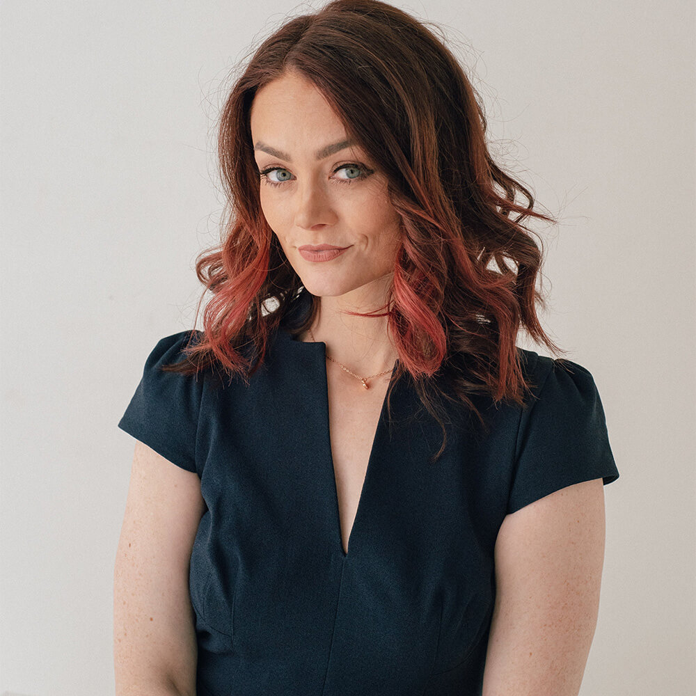Kristin Flanary smiling slightly, wearing a dark dress with a notched neckline and loose wavy hair with red highlights, standing against a plain light background.